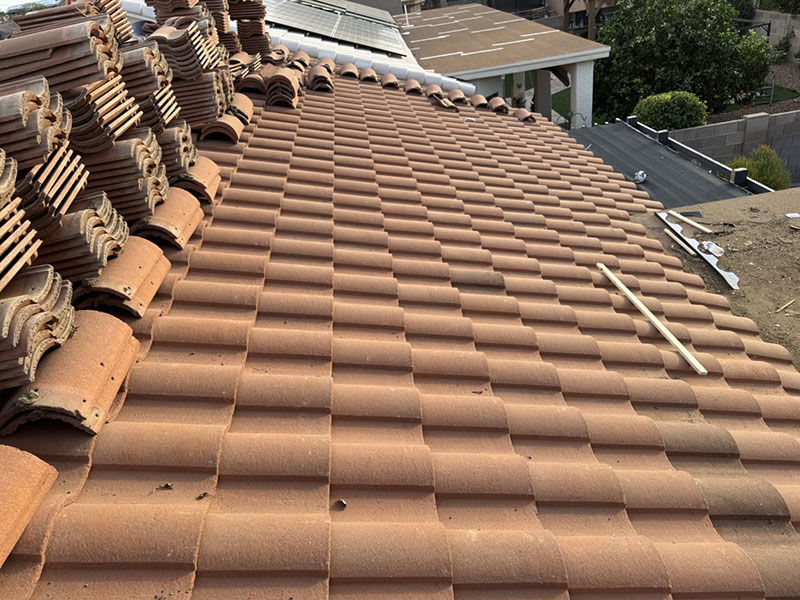 Stacks of terra-cotta roof tiles are placed on a partially completed tile roof. The tiles are arranged in neat rows, and construction materials are visible nearby. A street and trees can be seen in the background.