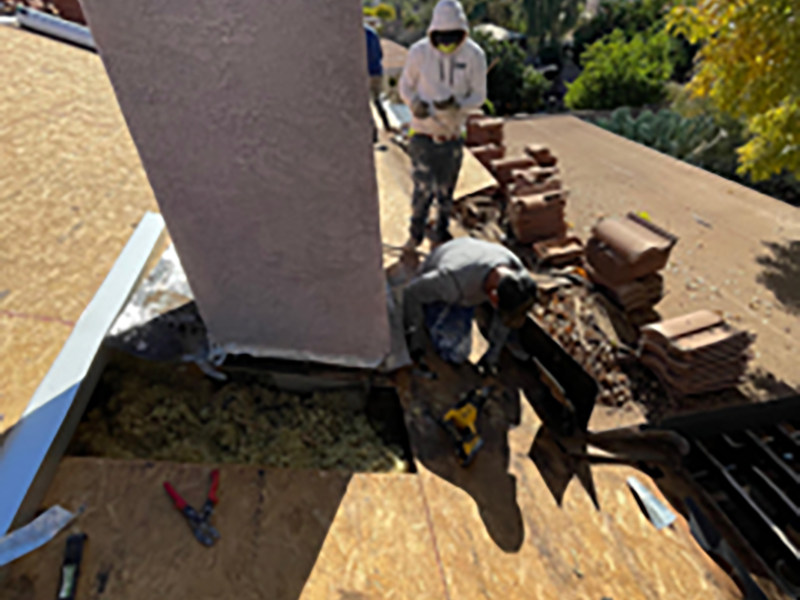 Two workers wearing protective gear repair a roof near a chimney, with tools and stacked tiles nearby. One worker kneels while the other stands, and part of the roofing is removed, exposing the underlying structure.