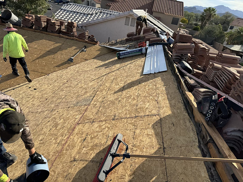 Workers are installing a new roof on a house, with piles of roof tiles and tools scattered around. Some workers are sweeping and others are handling materials on the wooden roof deck. Nearby homes and palm trees are visible.