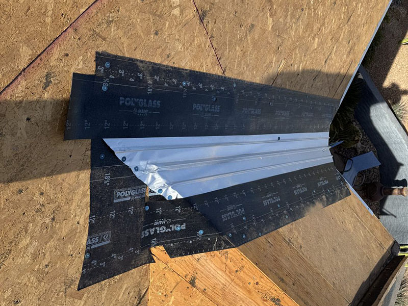 A close-up of a rooftop under construction showing a metal flashing installed over roofing underlayment, with wooden roof decking visible around the area.