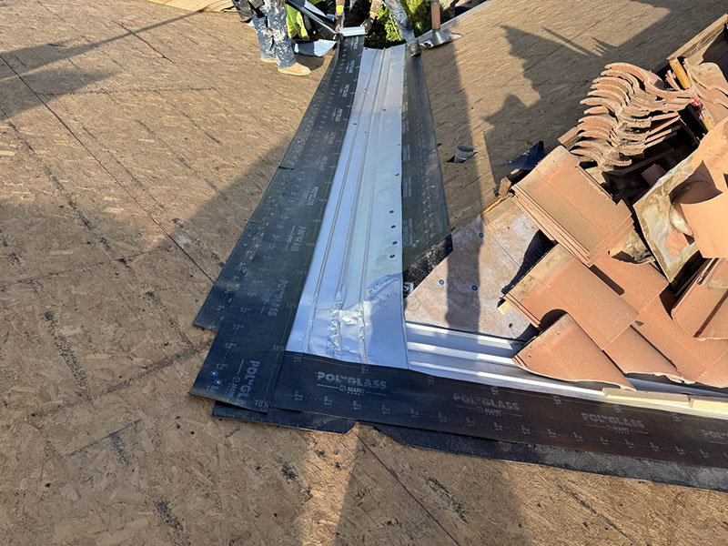 Close-up of a roof under construction, showing black waterproof membrane and metal flashing installed at a roof valley, with stacks of brown shingles or tiles nearby and part of a worker visible in the background.