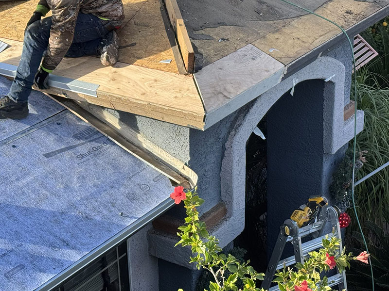 A worker in camo clothing repairs a corner section of a house roof, using plywood and tools. A ladder leans against the structure, and red hibiscus flowers are visible at the bottom right.