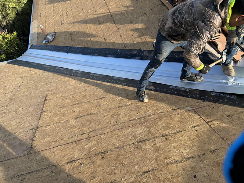 A person wearing a camouflage jacket and work gloves installs metal flashing on a sloped plywood roof under construction, with tools and materials visible nearby.
