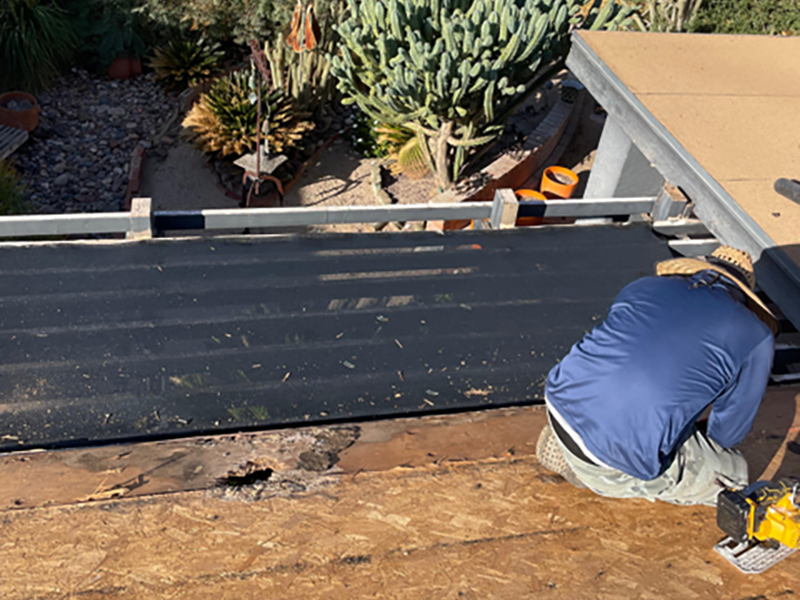 A person in a blue shirt and straw hat kneels on a roof, installing black roofing material, with tools nearby and a garden with cacti visible below.