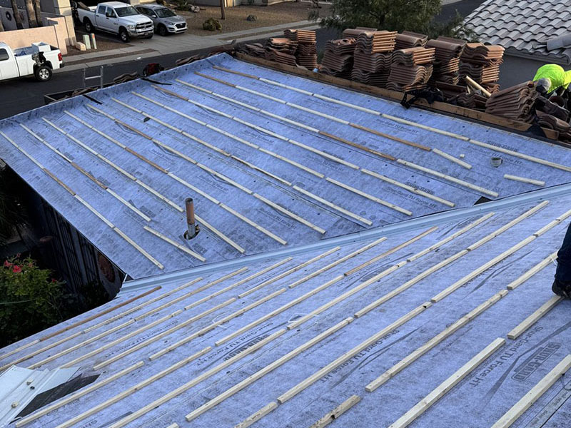 Roof under construction with underlayment and wooden battens installed. Stacks of tiles and a worker are visible on the upper part of the roof. Several vehicles are parked on the street below.