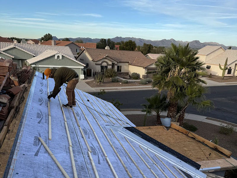 A worker wearing a yellow hard hat installs strips on a house roof under construction, with suburban homes, palm trees, and mountains in the background.