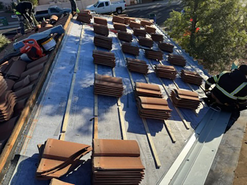 Rows of brown roofing tiles are neatly stacked on a rooftop, with a worker in safety gear kneeling on the right side and various tools visible. Trees and a street can be seen in the background.