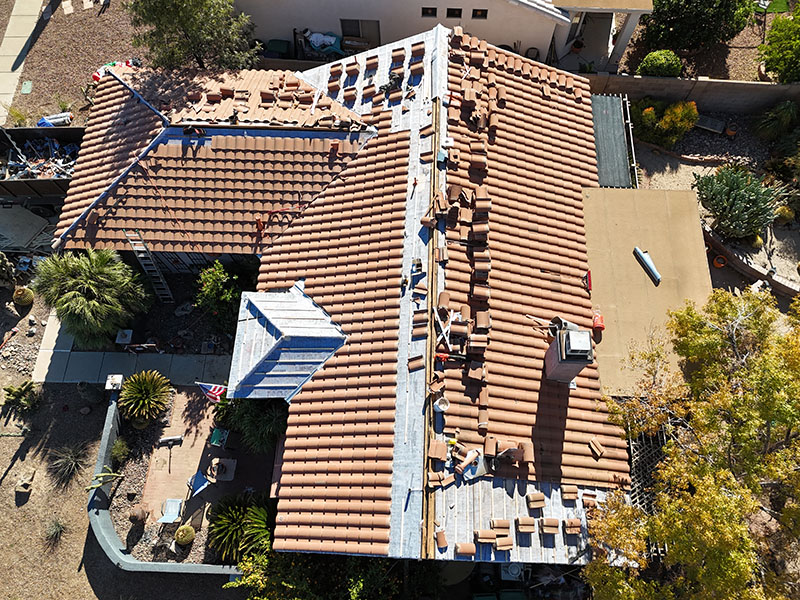 Aerial view of a house with workers replacing roof tiles; some sections have exposed underlayment, while others have new tiles partially installed. Tools and materials are visible on the roof and around the property.