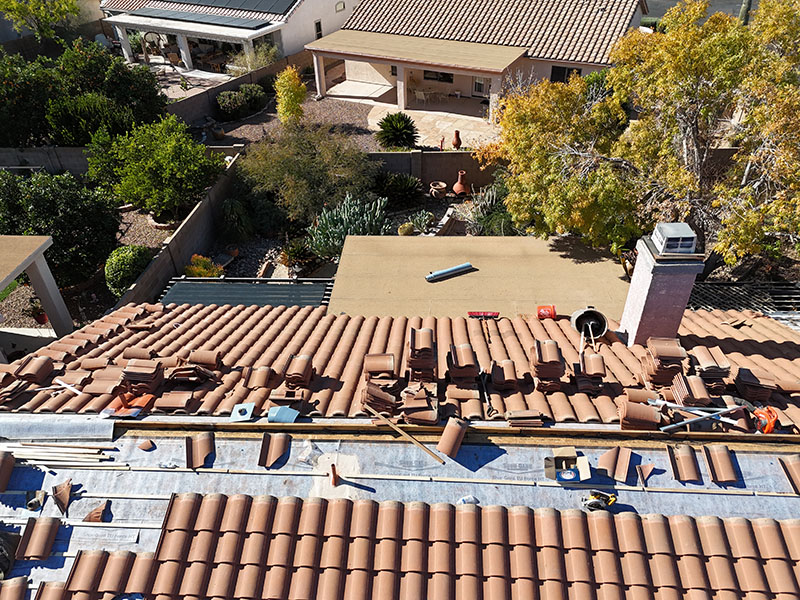 Aerial view of a house roof under repair with brown tiles, tools, and materials scattered across the surface. Nearby, there are trees, a chimney, and neighboring houses with landscaped backyards.