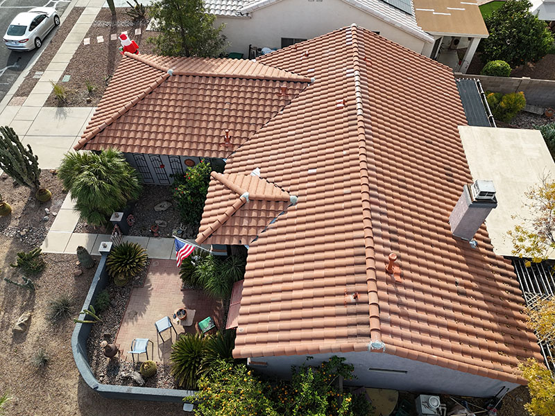 Aerial view of a house with a red tile roof, a small front patio with chairs and plants, American and Canadian flags displayed, and a desert-style yard with rocks and cacti.