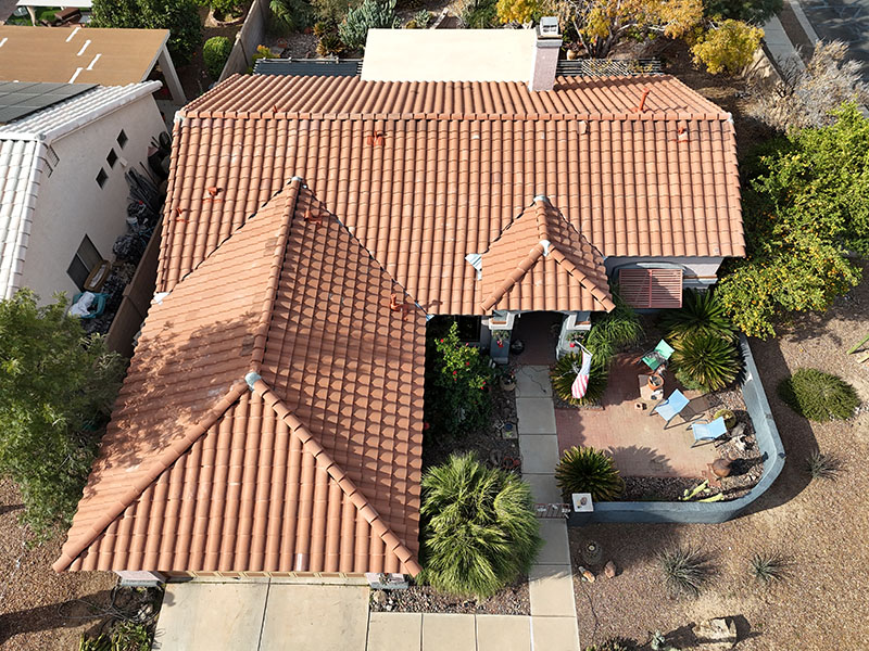 Aerial view of a single-story house with a red-tiled roof, surrounded by desert landscaping, small bushes, a concrete walkway, and a small patio area with outdoor seating and sun umbrellas.