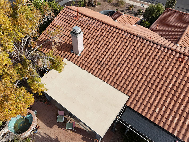 Aerial view of a house with a red tile roof, white chimney, beige patio cover, and a backyard area with patio furniture and a round hot tub surrounded by trees and plants.