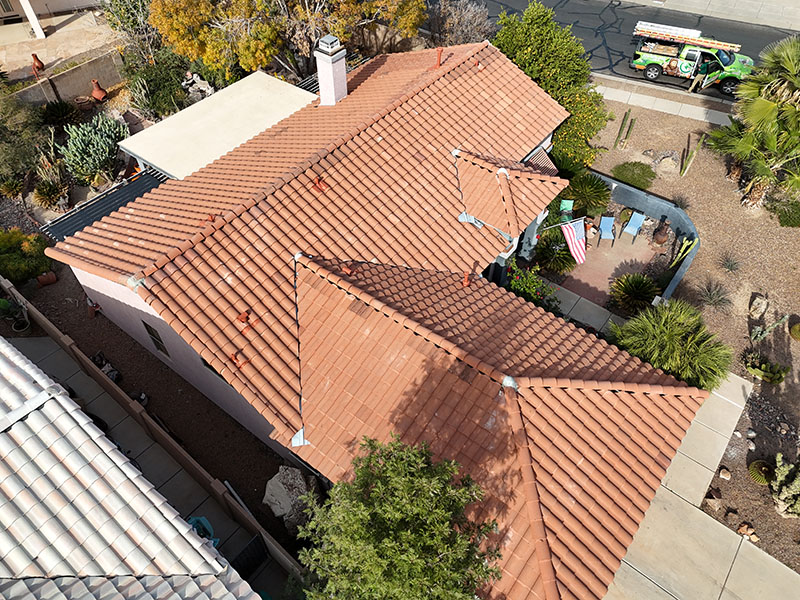 Aerial view of a house with a red tile roof, surrounded by desert landscaping, patio furniture, an American flag, and a tree near the entrance; a green utility truck is parked on the street.