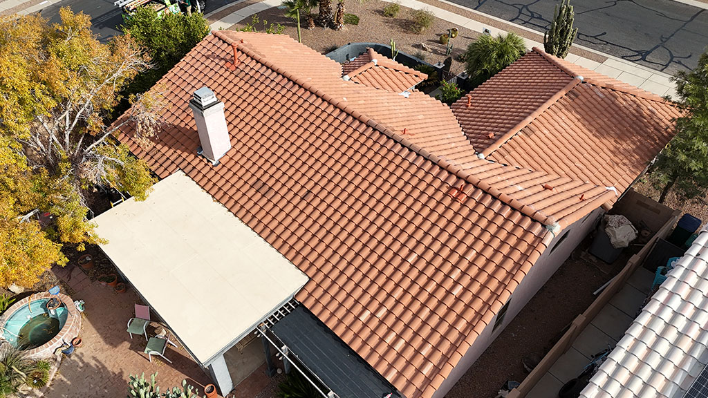 Aerial view of a house with a terracotta tile roof, a white chimney, a small backyard with a round hot tub, trees, and surrounding streets. The property has a patio area and landscaping with rocks and desert plants.