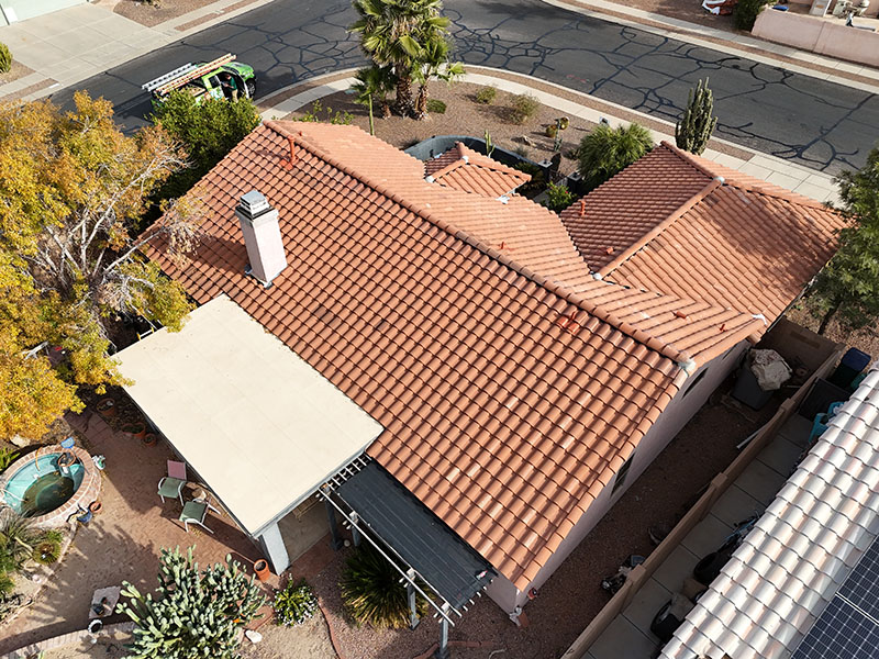 Aerial view of a house with red-tiled roof, surrounded by trees and desert landscaping, next to a street with cracked pavement and a green truck parked nearby.