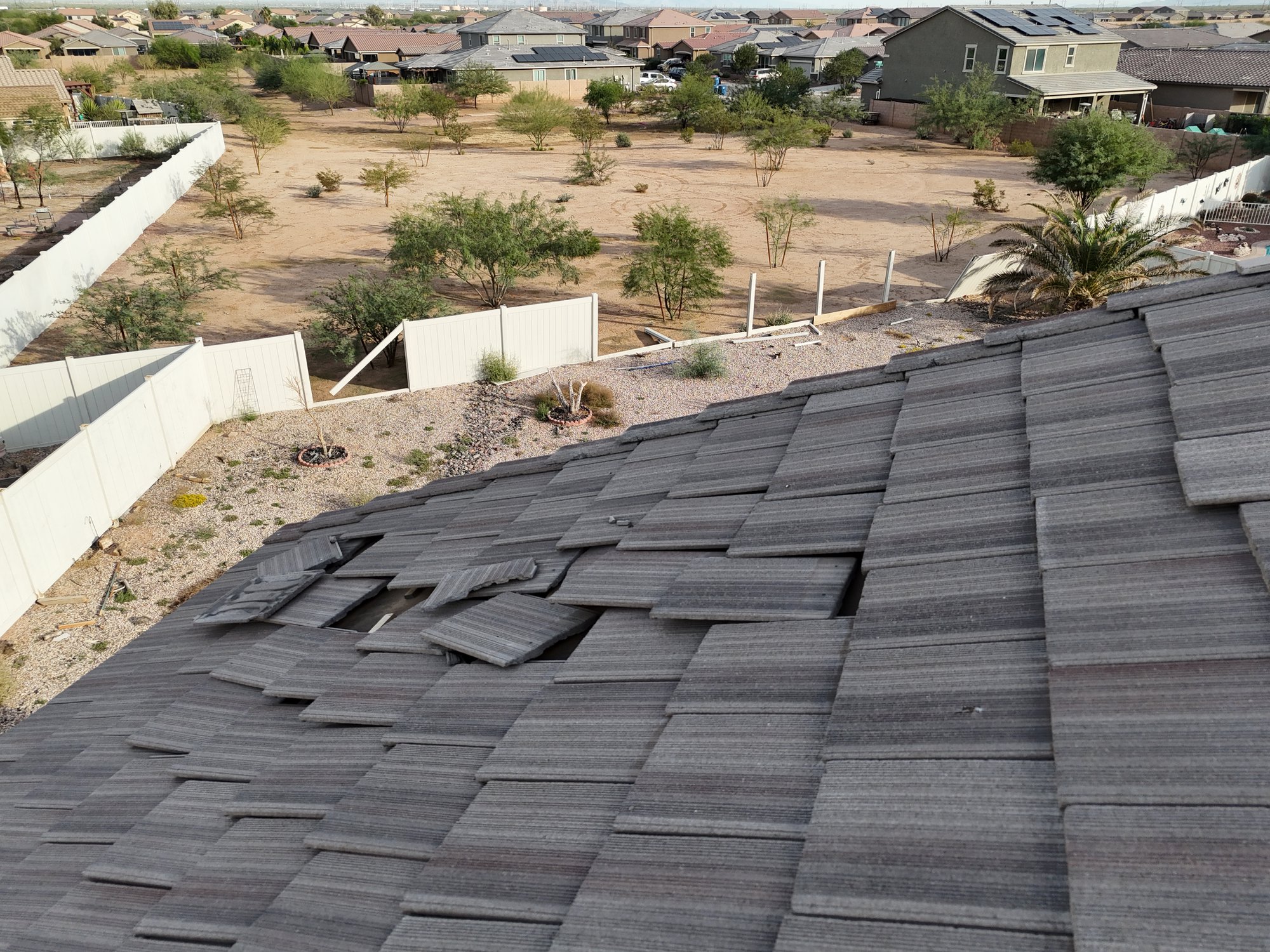 A rooftop with several damaged and missing shingles is shown in the foreground, with a fenced backyard, sparse trees, and neighboring houses visible in the background. default